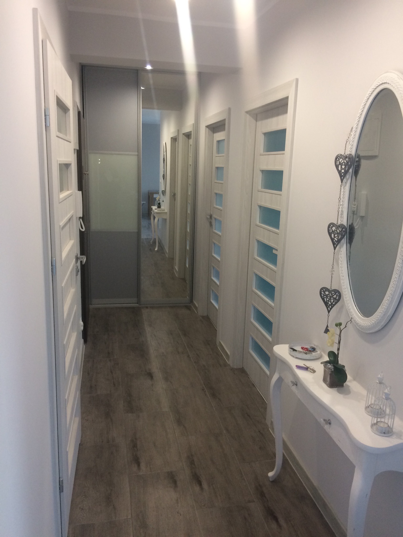 Jasny korytarz z drewnopodobną podłogą, białymi drzwiami z przeszkleniami, lustrem w ozdobnej ramie i konsolą z dekoracjami.