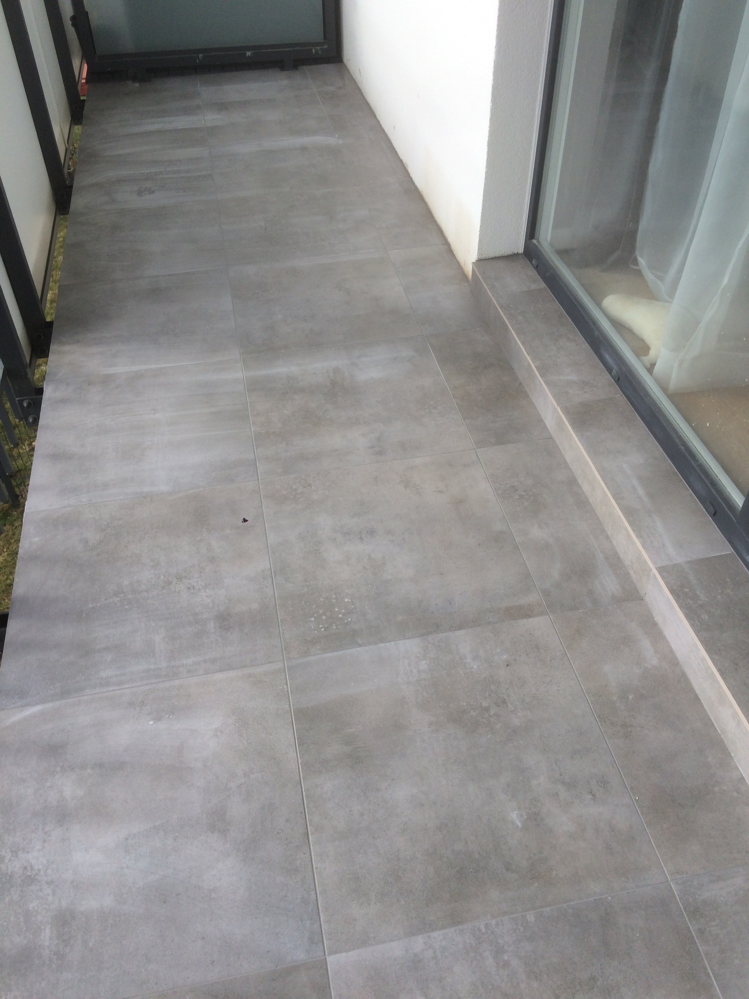 Wyłożony szarymi płytkami balkon z widocznym oknem balkonowym i metalową barierką.