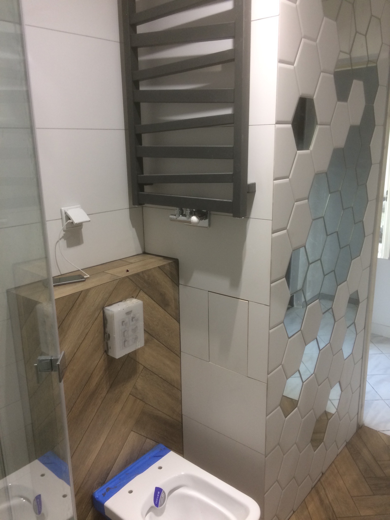 Nowoczesna łazienka z heksagonalnymi płytkami z lustrzanymi wstawkami, drewnopodobną zabudową WC, grzejnikiem drabinkowym i fragmentem kabiny prysznicowej.