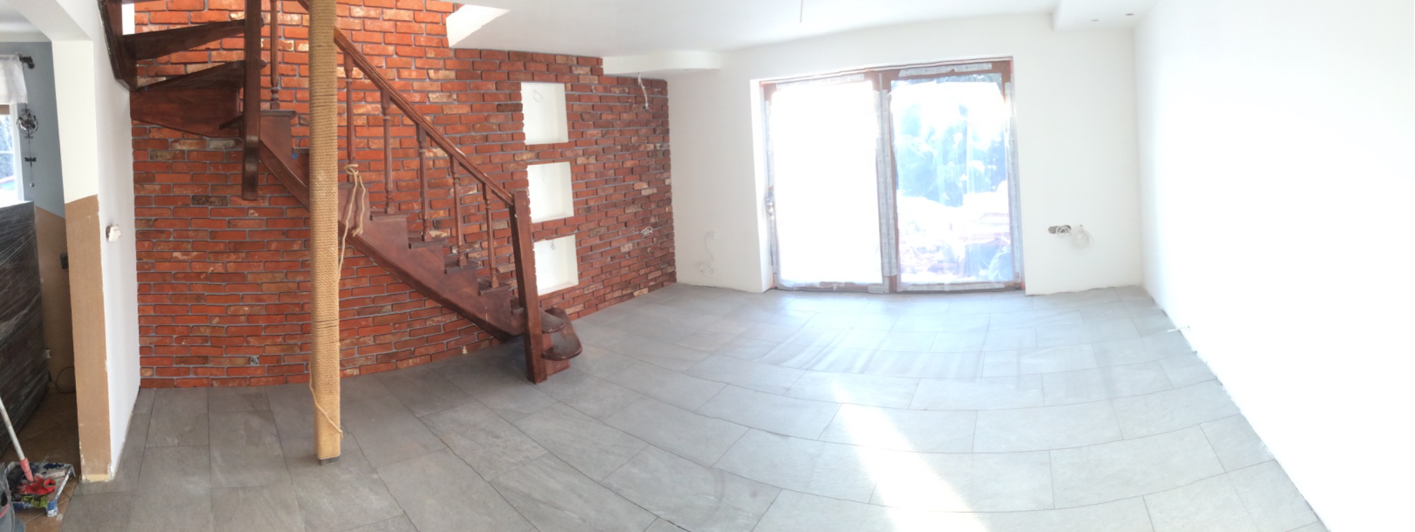 Panoramiczne ujęcie wnętrza w trakcie wykończenia: ceglana ściana z wnękami, drewniane schody spiralne z liną, szara podłoga z płytek, okno balkonowe z folią ochronną, białe ściany w surowym stanie.