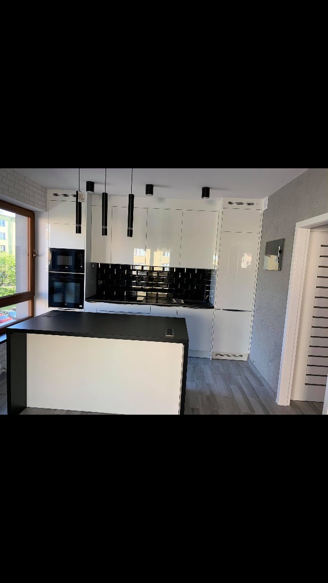 Nowoczesna kuchnia z białą zabudową, czarnymi akcentami i wyspą kuchenną, widoczna przez okno z ceglanym obramowaniem, oświetlona wiszącymi lampami.