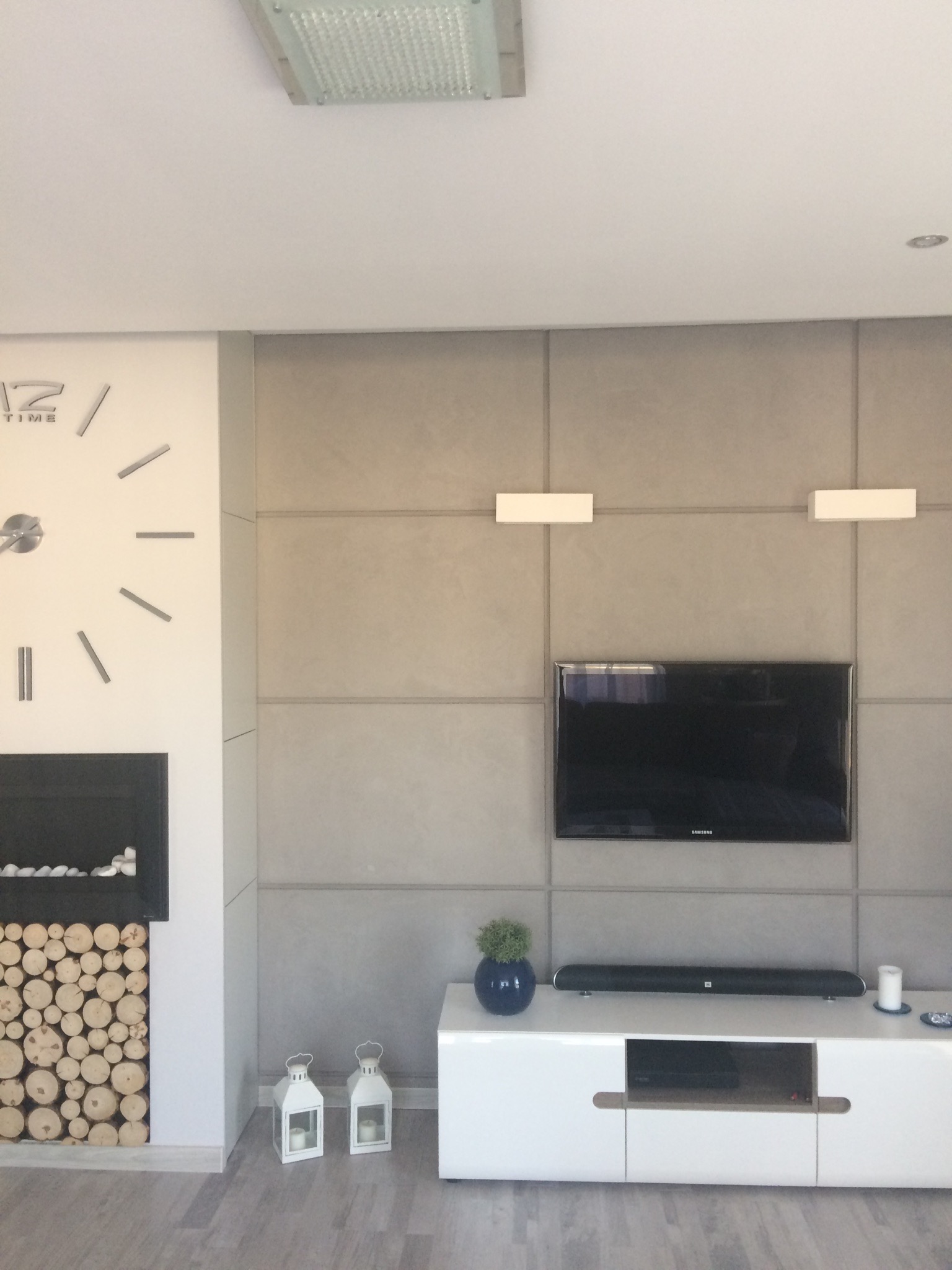 Nowoczesny salon z szarą ścianą panelową, telewizorem, kominkiem z drewnem, białymi lampionami i zegarem ściennym.