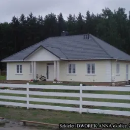 Gotowy dom szkieletowy z szarą dach&oacute;wką, białymi oknami i kolumnami werandy, ogrodzony białym płotem, z rowerem opartym o ścianę.