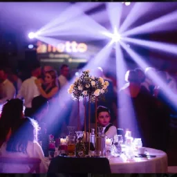 Sala balowa z oświetleniem scenicznym, stołami z dekoracjami kwiatowymi i gośćmi w tle.