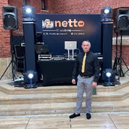 DJ w czarnej koszuli i żółtym krawacie pozuje na tle sprzętu: mikser, laptop, głośniki i oświetlenie na scenie z ceglaną ścianą w tle.