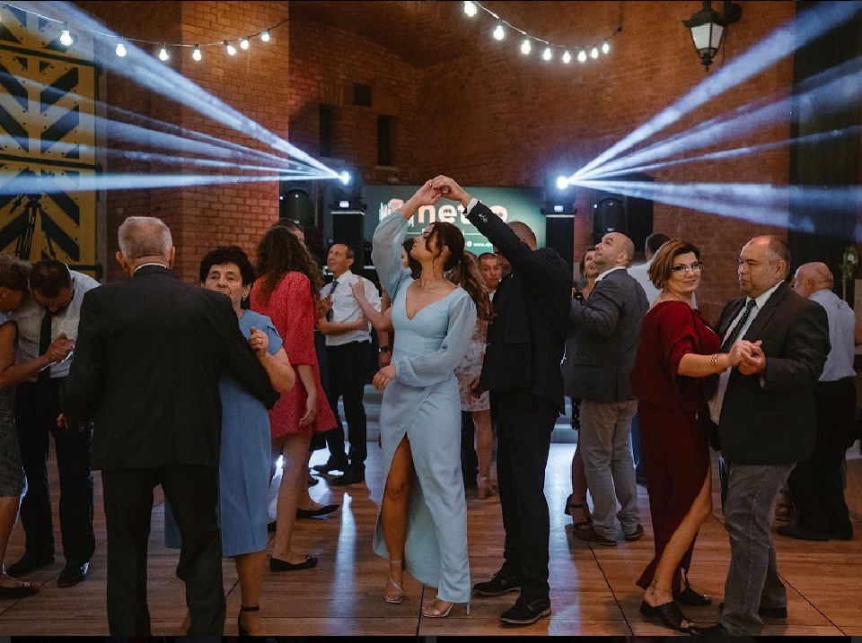 Para młoda tańczy na parkiecie, oświetlona reflektorami podczas uroczystości weselnej w ceglanym wnętrzu z wiszącymi żarówkami.