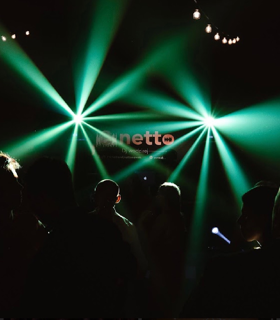 Zielone reflektory oświetlają sylwetki publiczności podczas wydarzenia z DJ-em, widoczne żarówki w tle.