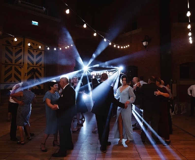 Para tańczy na parkiecie podczas wesela, oświetlona reflektorami i girlandami żarówek, w tle ceglana ściana i dekoracyjne drzwi stodoły.