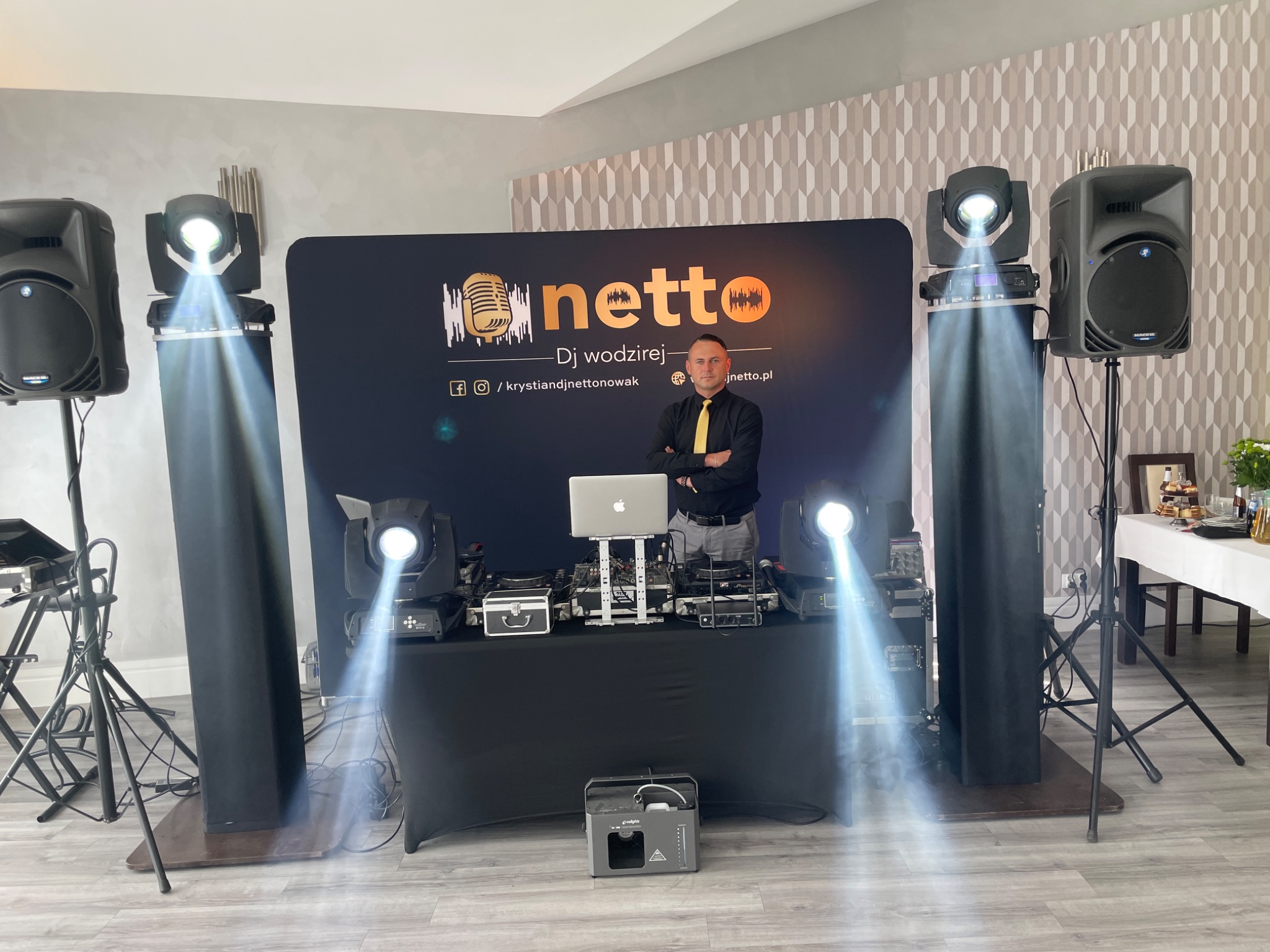 DJ wodzirej w czarnej koszuli i żółtym krawacie stoi za konsoletą z laptopem, otoczony profesjonalnym nagłośnieniem i oświetleniem z logiem firmy na tle.