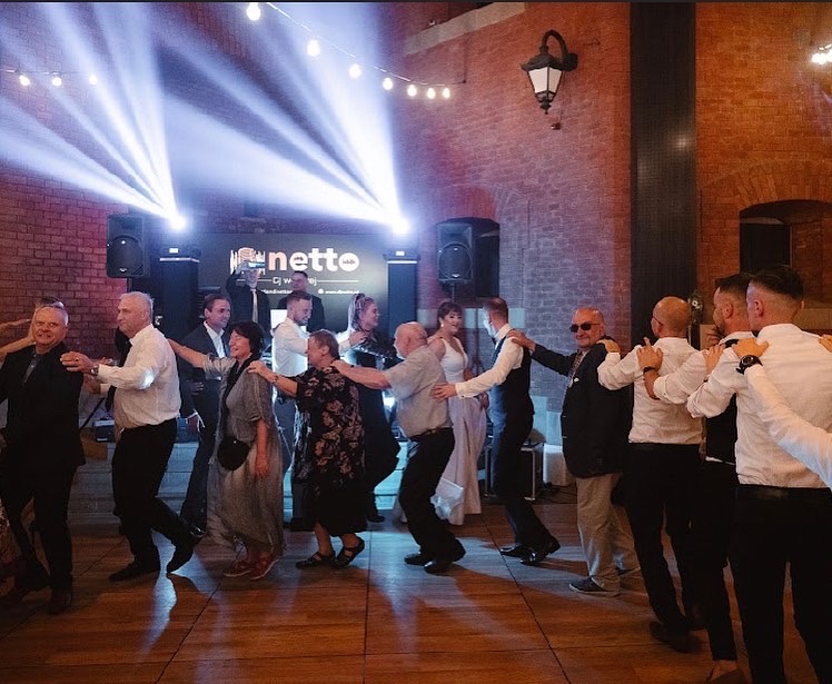 Rozbawieni goście w trakcie tańca korowodowego na parkiecie w Bochni, oświetlonym przez reflektory i nagłośnionym przez profesjonalny sprzęt. W tle widoczna ceglana ściana i dekoracyjne lampki.
