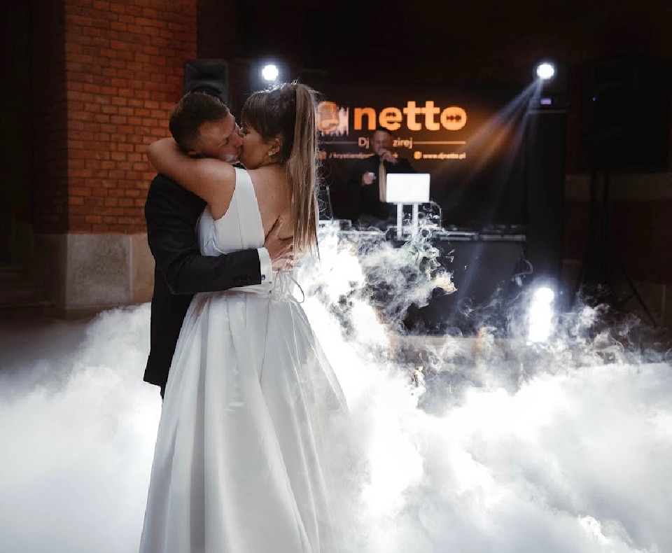Pocałunek nowożeńców na tle sceny z DJ-em, oświetleniem i ciężkim dymem podczas wesela w ceglanym budynku.