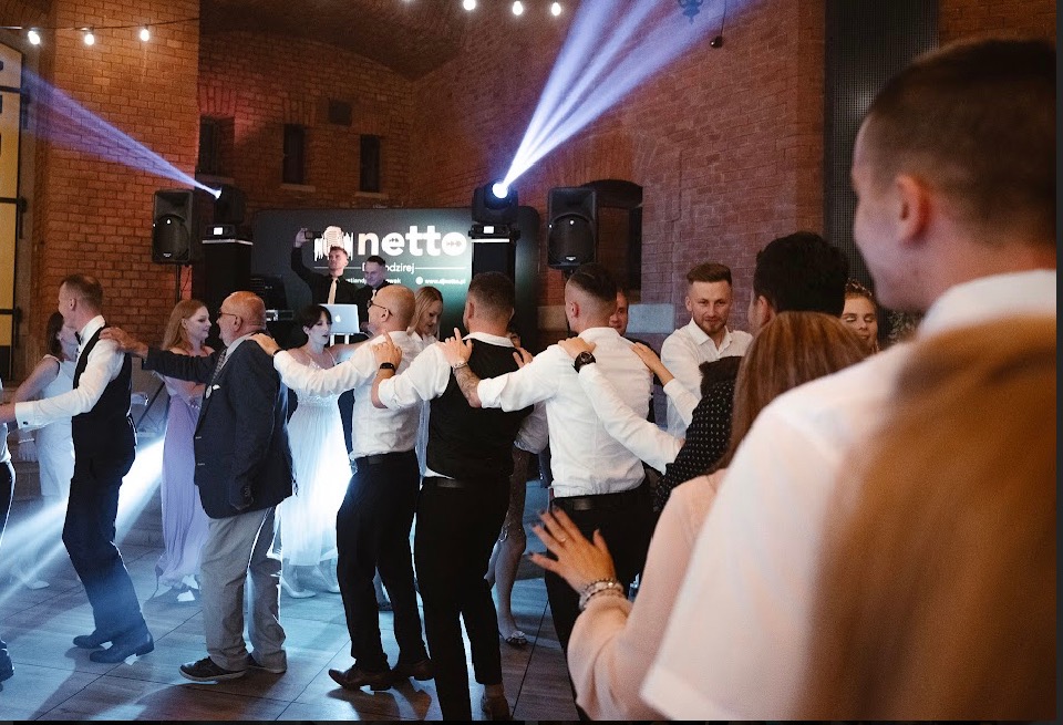 Goście weselni tańczą w kręgu przy muzyce DJ'a z podświetleniem scenicznym w ceglanym wnętrzu sali weselnej.
