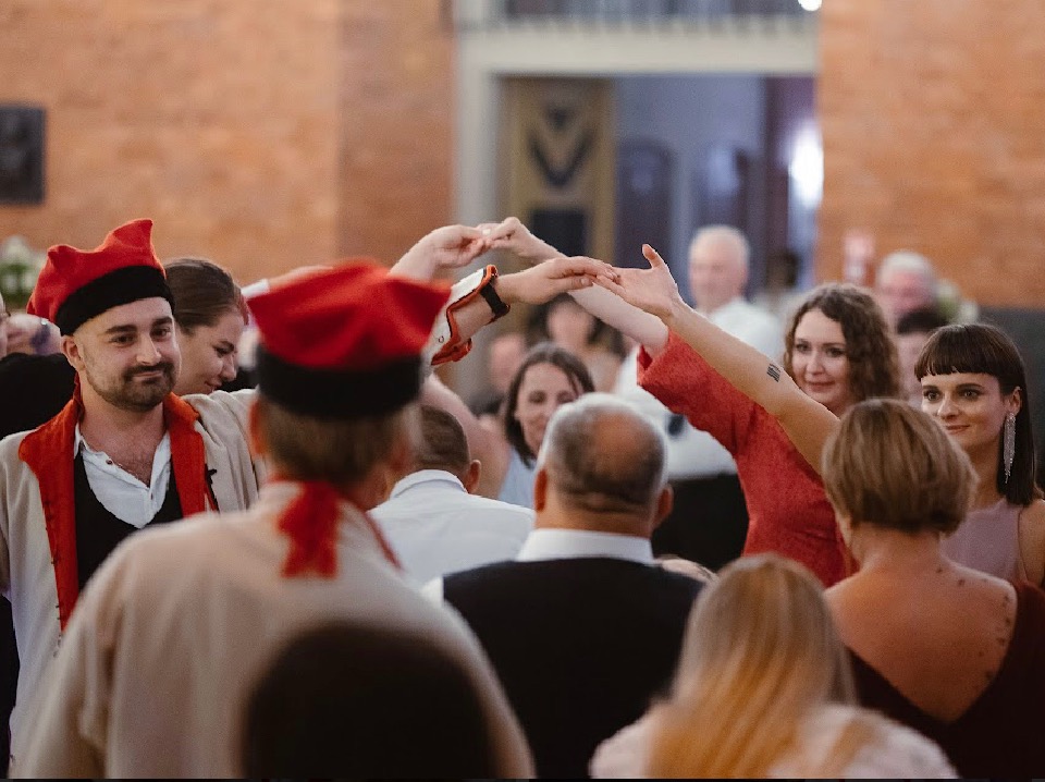 Uczestnicy wesela tworzą tunel z rąk, mężczyzna w stroju ludowym z czerwonym czepcem uśmiecha się.