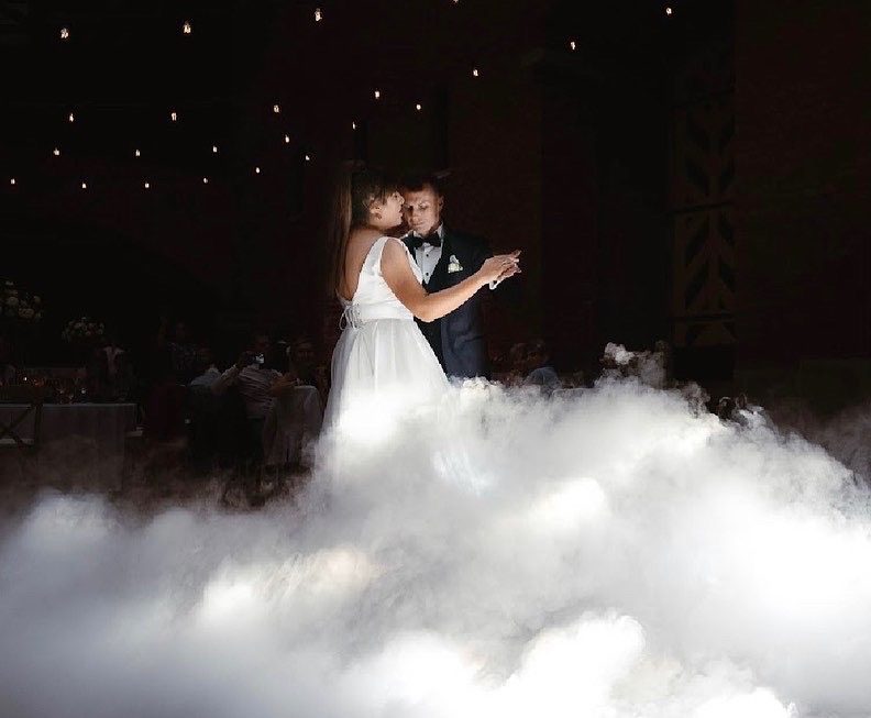 Para młoda tańczy pierwszy taniec w chmurze dymu na sali weselnej z dekoracyjnym oświetleniem.