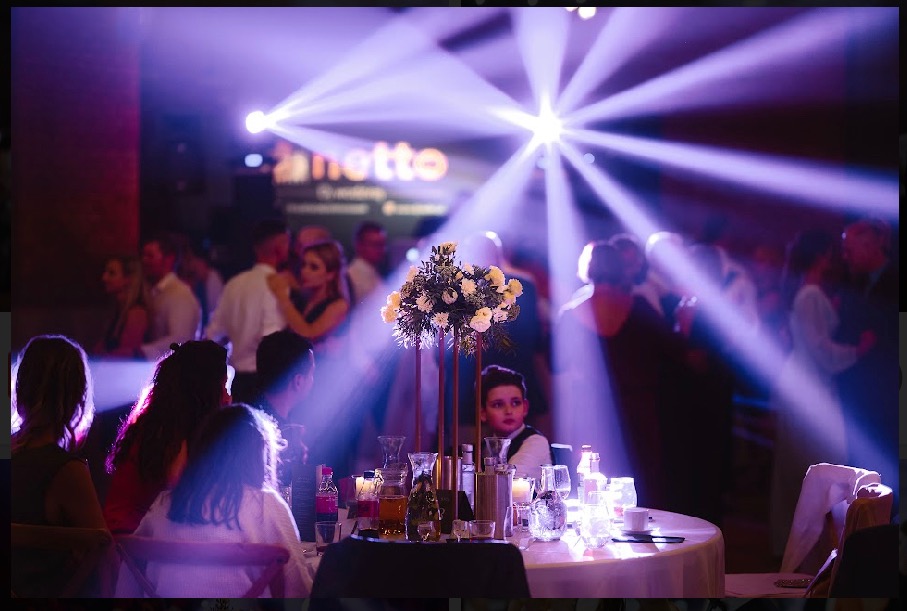 Sala balowa z oświetleniem scenicznym, stołami z dekoracjami kwiatowymi i gośćmi w tle.