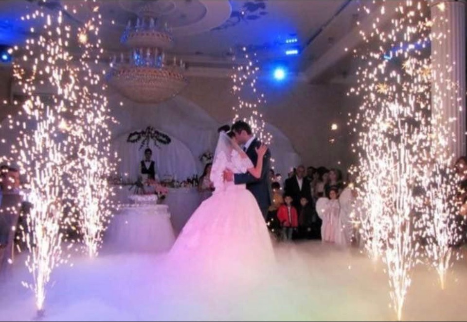 Para młoda tańczy w chmurze dymu otoczona fontannami iskier na sali balowej z kryształowym żyrandolem i gośćmi w tle.