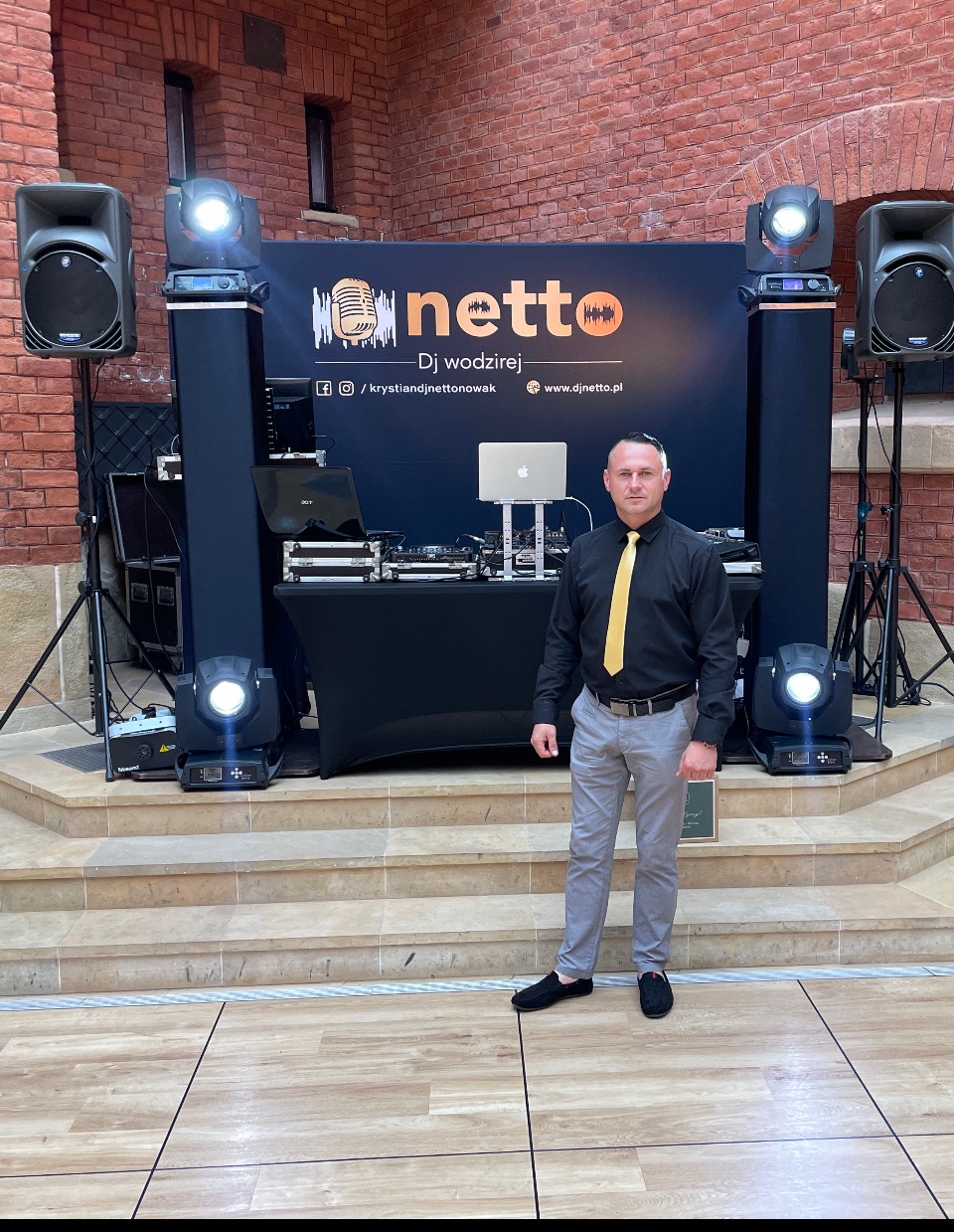 DJ w czarnej koszuli i żółtym krawacie pozuje na tle sprzętu: mikser, laptop, głośniki i oświetlenie na scenie z ceglaną ścianą w tle.