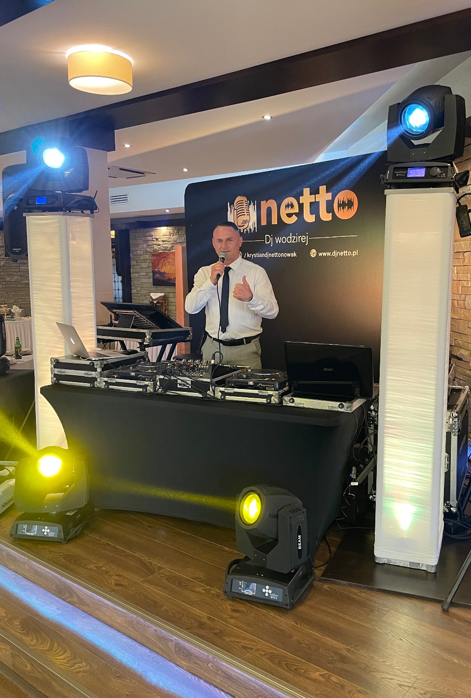 DJ z mikrofonem i uniesionym kciukiem na tle stanowiska z mikserem, laptopami i oświetleniem scenicznym podczas imprezy.