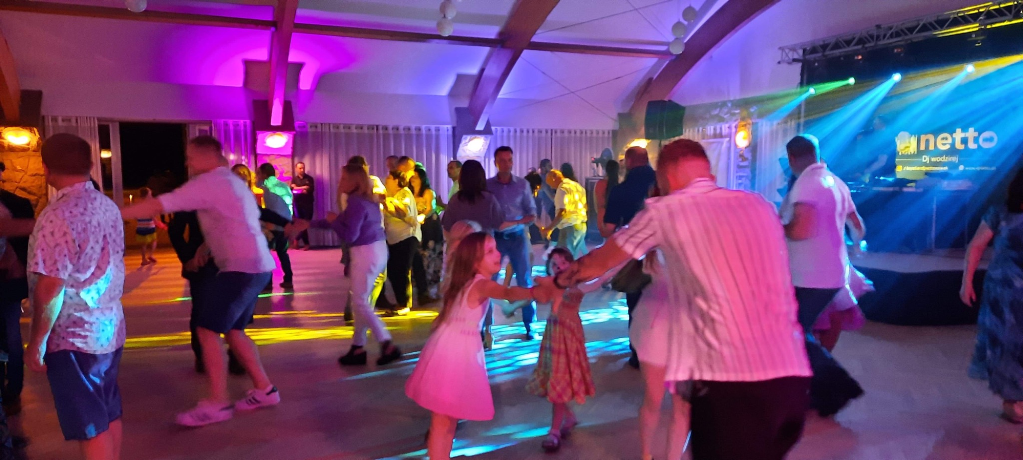 Parkiet taneczny wypełniony tańczącymi osobami, oświetlony kolorowymi światłami, z widoczną sceną w tle i logiem DJ-a.