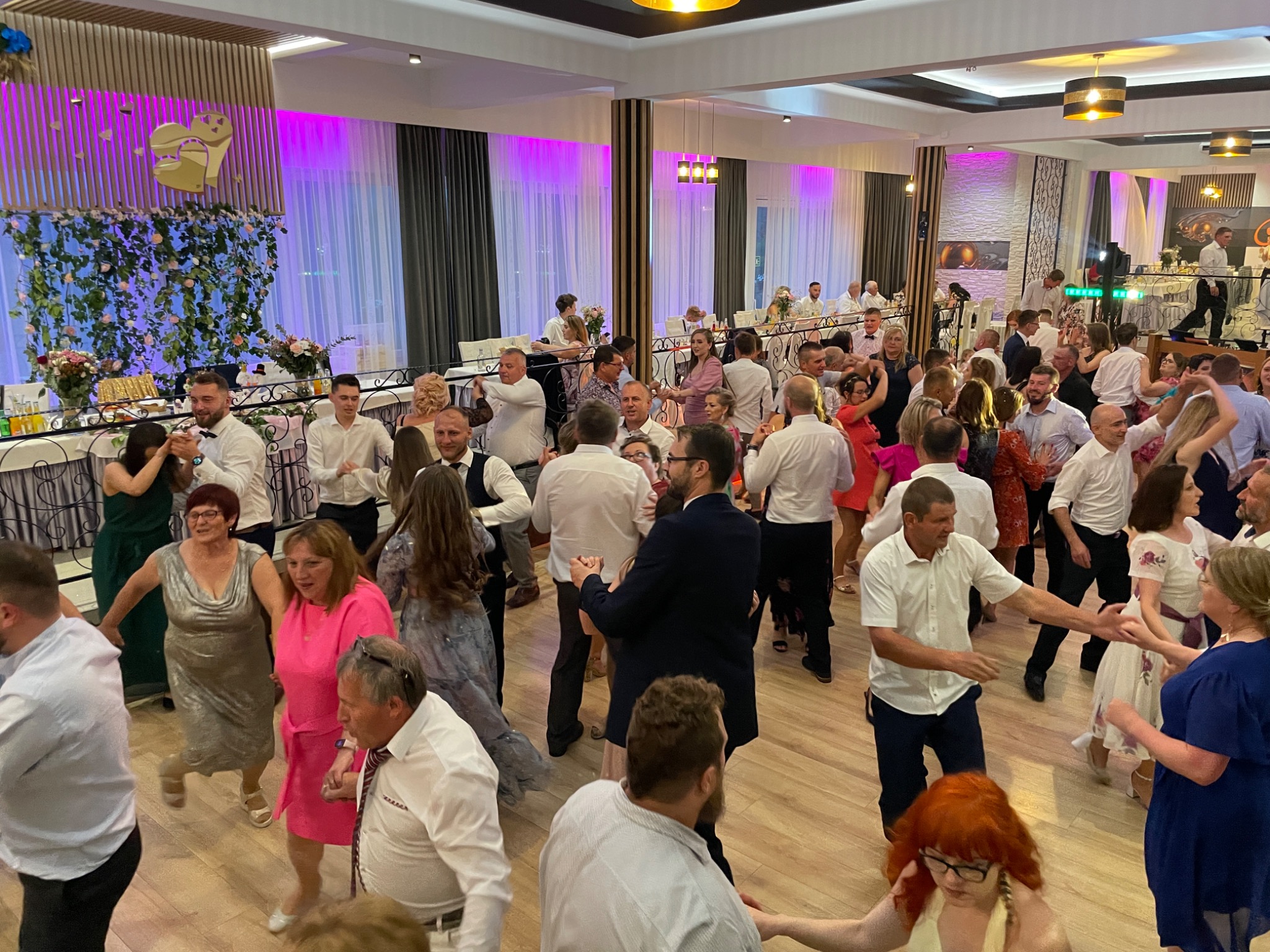 Parkiet taneczny wypełniony gośćmi weselnymi podczas zabawy, dekoracje kwiatowe i oświetlenie sceniczne w tle.