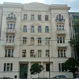 Fasada odnowionej, jasnej kamienicy z balkonami i dekoracyjnymi elementami architektonicznymi, widoczna z zewnątrz, z drzewami wzdłuż ulicy.