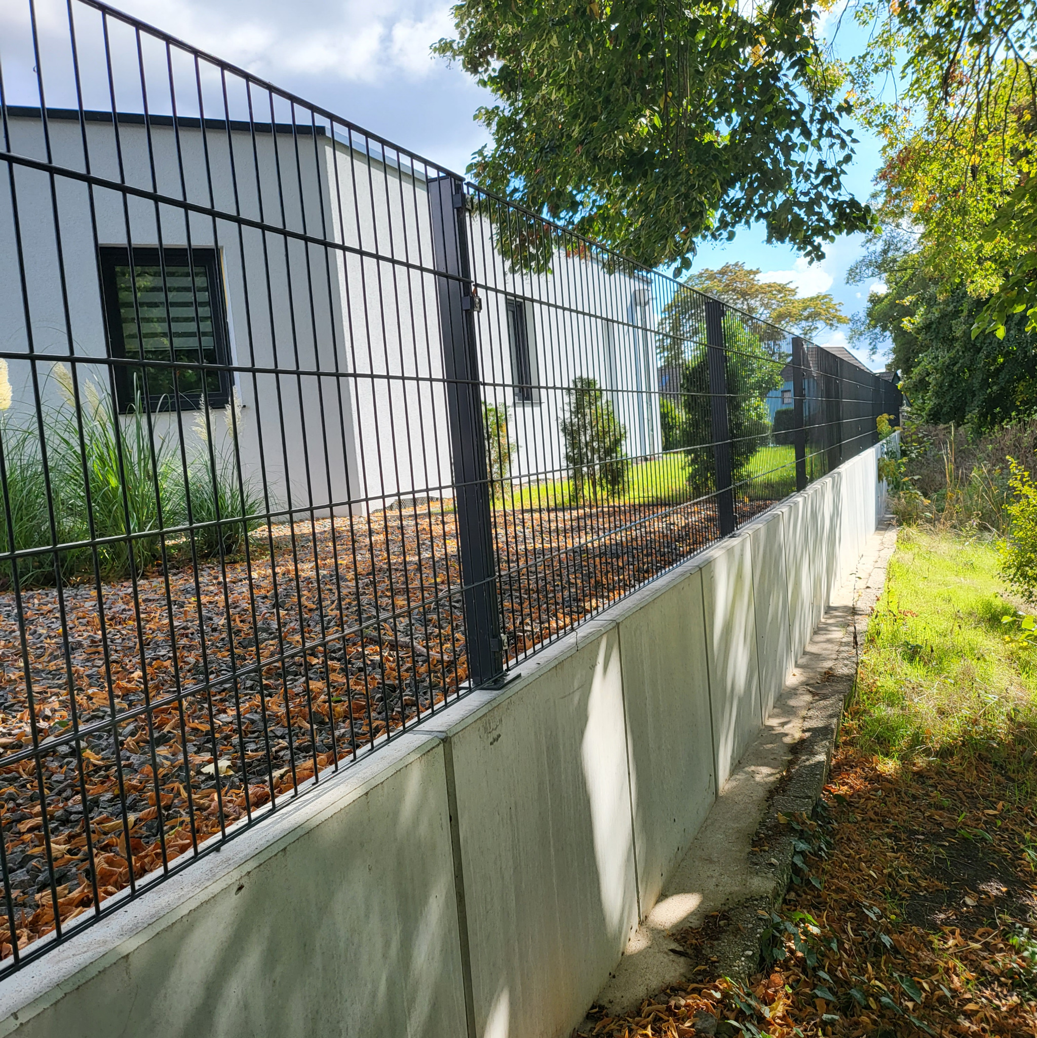 Czarny panelowy system ogrodzeniowy na betonowej podmurówce w Gubinie, widok z boku, z liśćmi i kamieniami pod ogrodzeniem.