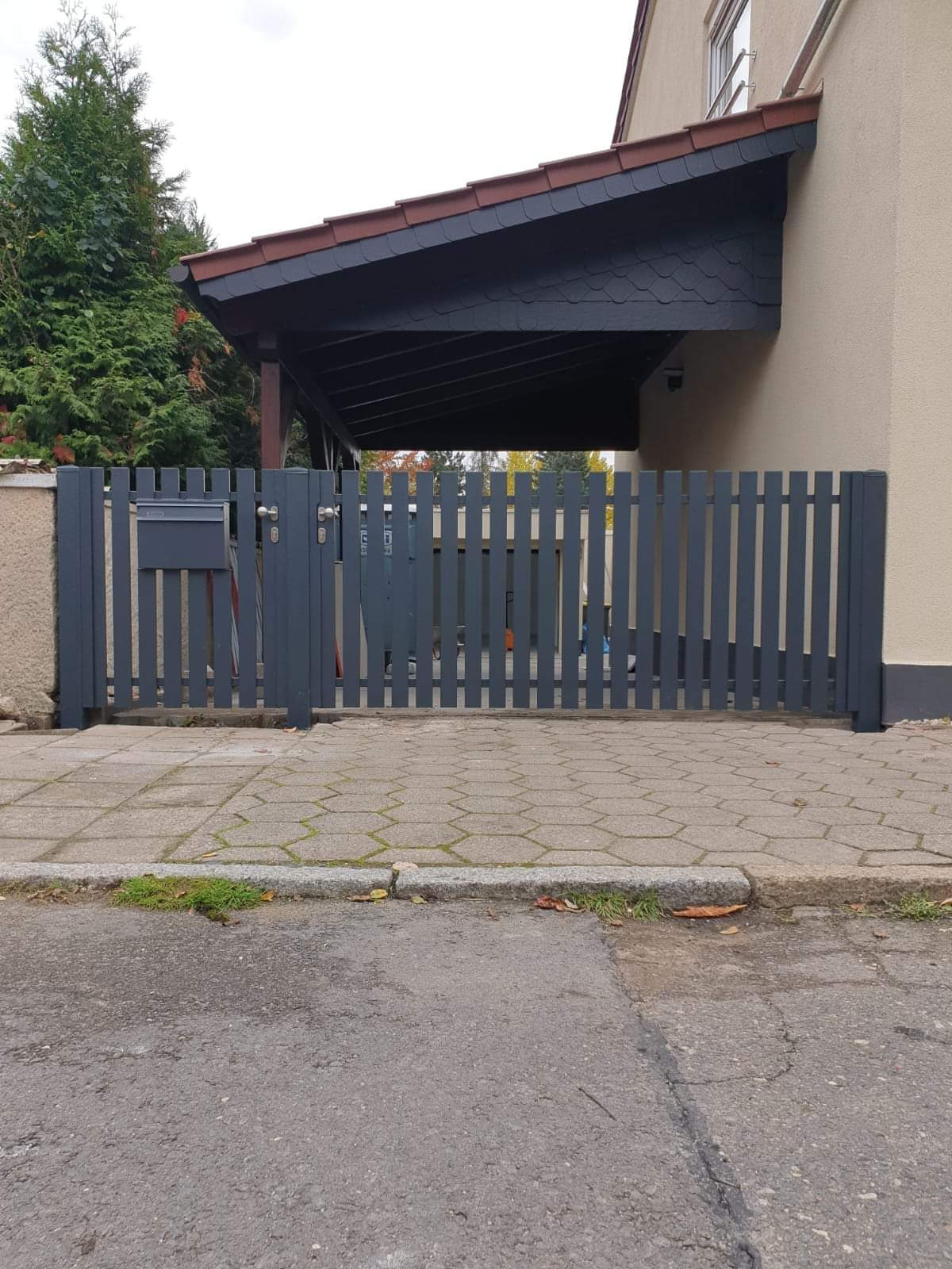 Nowoczesna, szara brama wjazdowa z pionowych paneli zintegrowana ze skrzynką na listy i furtką, na tle domu z zadaszonym podjazdem.