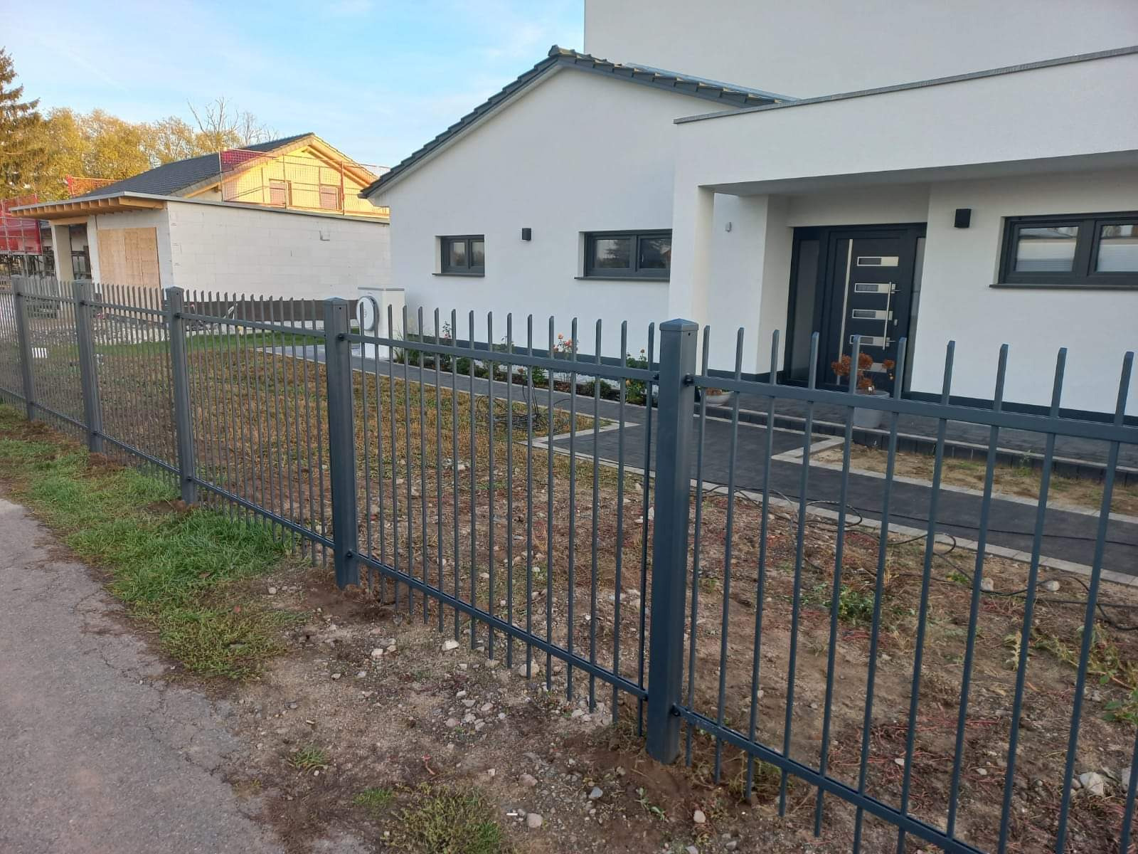 Szare metalowe ogrodzenie palisadowe przed nowoczesnym domem z białymi ścianami i szarymi oknami, widoczne trawnik i ścieżka z kostki.