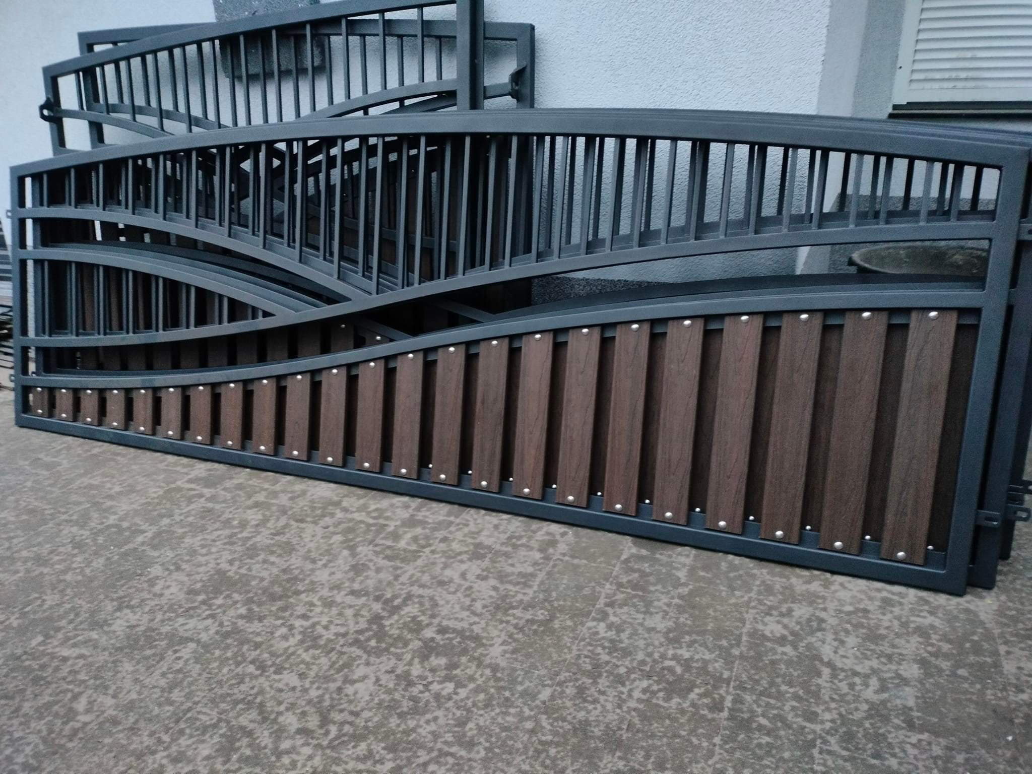 Elementy bramy wjazdowej: metalowa rama w kolorze grafitowym z dekoracyjnym, falistym wypełnieniem z brązowych paneli, widoczne detale połączeń nitowych.
