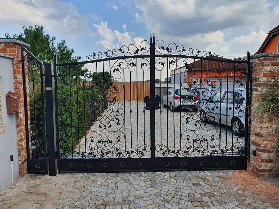 Czarna, ozdobna brama wjazdowa z kutego żelaza, z dekoracyjnymi elementami roślinnymi, zamontowana pomiędzy dwoma murowanymi słupkami na posesji z brukowaną nawierzchnią, widoczne dwa samochody...
