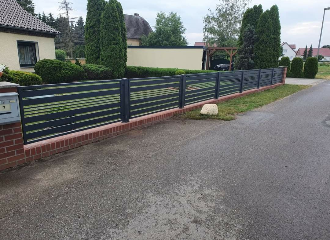 Nowoczesne ogrodzenie metalowe z poziomymi prętami, osadzone na niskim murku z czerwonej cegły, widok z ulicy, zieleń w tle.