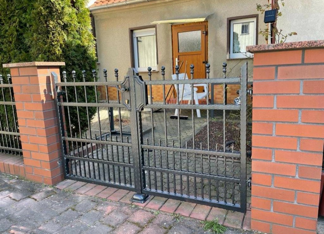 Metalowa brama wjazdowa z dekoracyjnymi elementami, osadzona między murowanymi słupkami z czerwonej cegły, na tle domu z drewnianymi drzwiami i roślinnością.