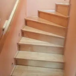 Jasne, drewniane schody z poręczą, oświetlone małymi punktowymi lampami w ścianie, prowadzące w górę.