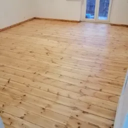 Jasna, odnowiona podłoga z desek w pokoju z białymi ścianami i oknem balkonowym.