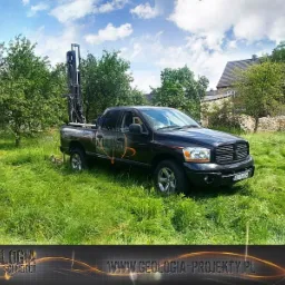 Czarny pickup z wiertnicą na pace, stojący na trawiastym terenie w słoneczny dzień, widoczne drzewa i budynek w tle.