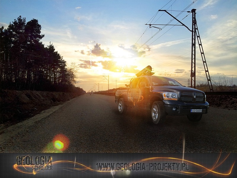 Czarny pickup z zamontowanym sprzętem pomiarowym na pace, stojący na żwirowej drodze w pobliżu torów kolejowych, z zachodzącym słońcem w tle.