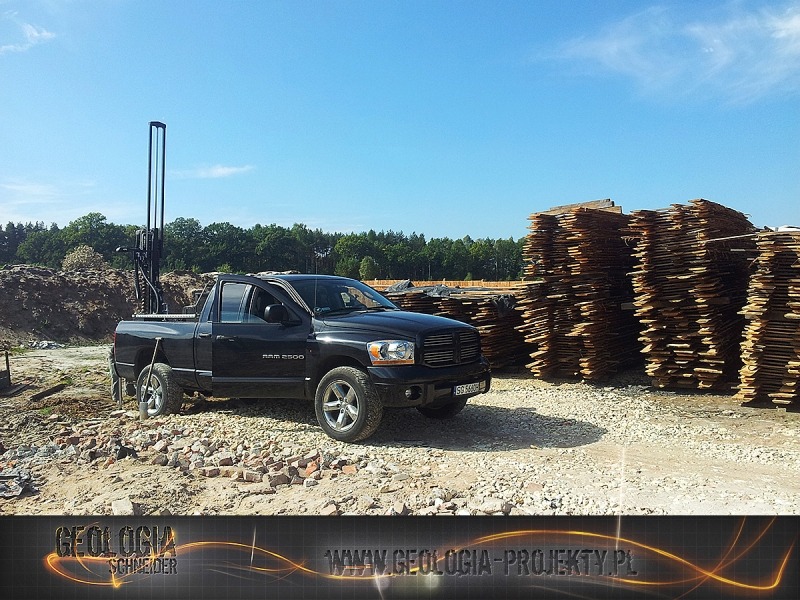 Czarny pickup Dodge Ram 2500 z wiertnicą w przestrzeni ładunkowej na placu budowy, w tle stosy desek i las.
