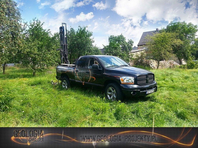 Czarny pickup z wiertnicą na pace, stojący na trawiastym terenie w słoneczny dzień, widoczne drzewa i budynek w tle.