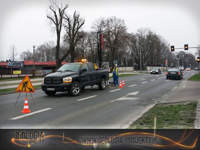 Czarny pickup z wiertnicą na środku ruchliwej ulicy, oznaczony trójkątnym znakiem ostrzegawczym i pomarańczowymi pachołkami; pracownik w odblaskowej kamizelce obok wiertnicy.