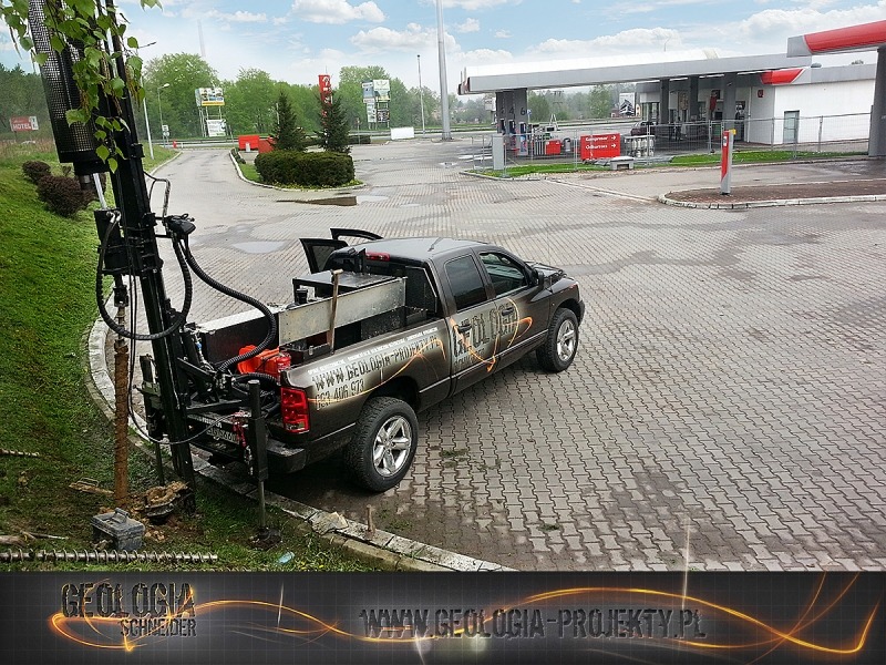 Wiertnica zamontowana na pick-upie Dodge Ram z logo firmy, wiercąca otwór w ziemi na tle stacji benzynowej.