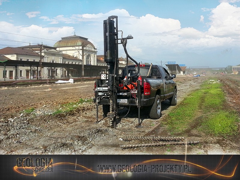 Wiertnica zamontowana na samochodzie terenowym, przygotowana do wiercenia gruntu na terenie budowy w pobliżu torów kolejowych i budynku dworca.