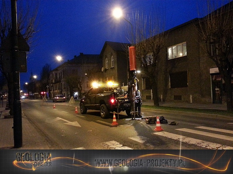 Prace terenowe z użyciem wiertnicy zamontowanej na pick-upie, oświetlonej lampami roboczymi, na tle zabudowy miejskiej o zmierzchu.