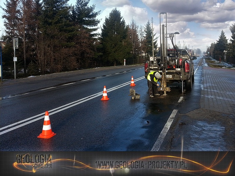 Pracownik w kamizelce odblaskowej obsługuje wiertnicę zamontowaną na pick-upie na mokrej jezdni, wygrodzonej pomarańczowymi pachołkami.