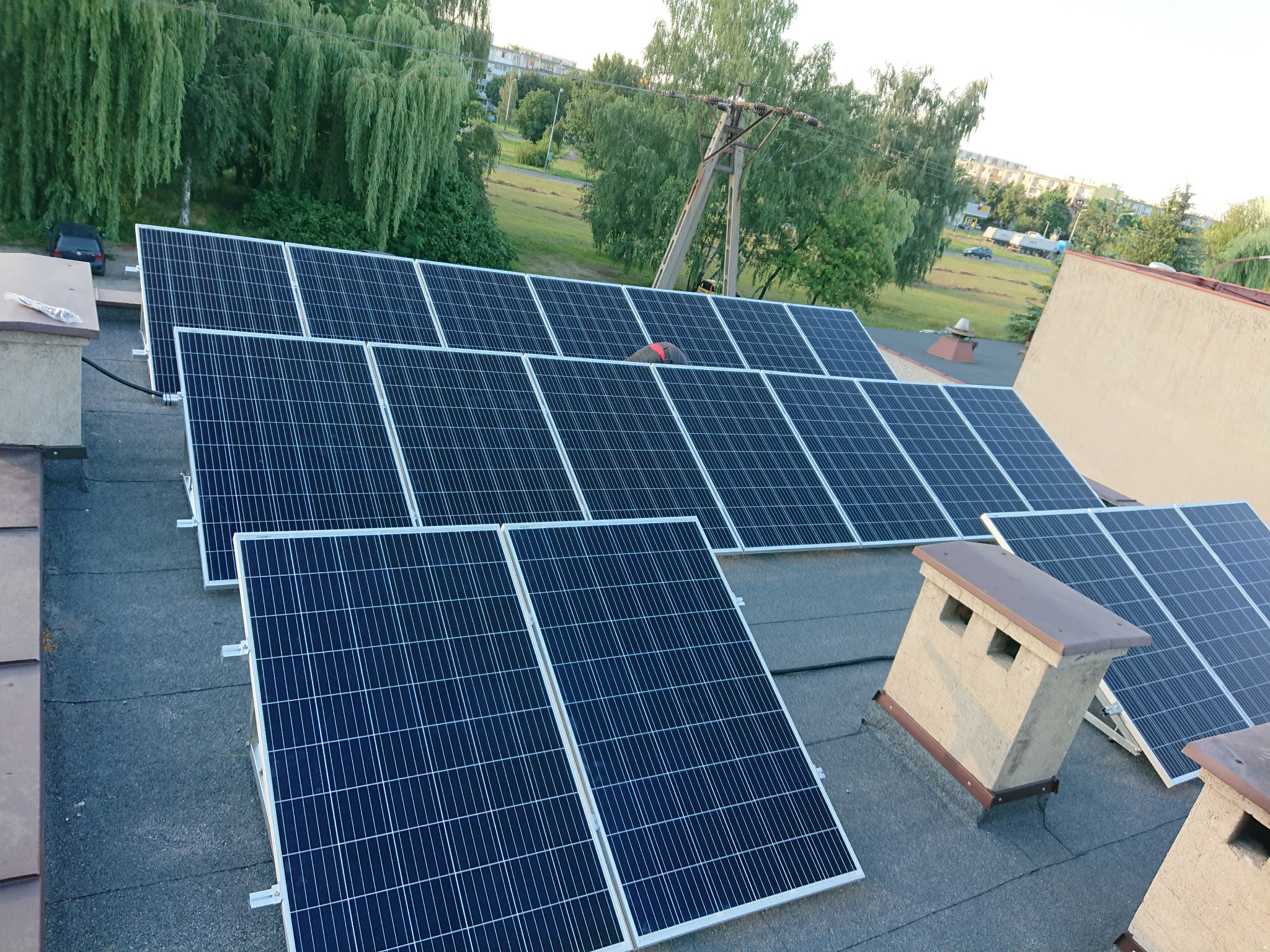 Instalacja fotowoltaiczna na dachu budynku z kominami wentylacyjnymi, widok z góry na panele i otoczenie z zielenią i zabudowaniami w tle.