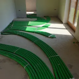 Zielone rury ogrzewania podłogowego ułożone w pętle na surowym podłożu w trakcie instalacji w jasnym pomieszczeniu z oknem.