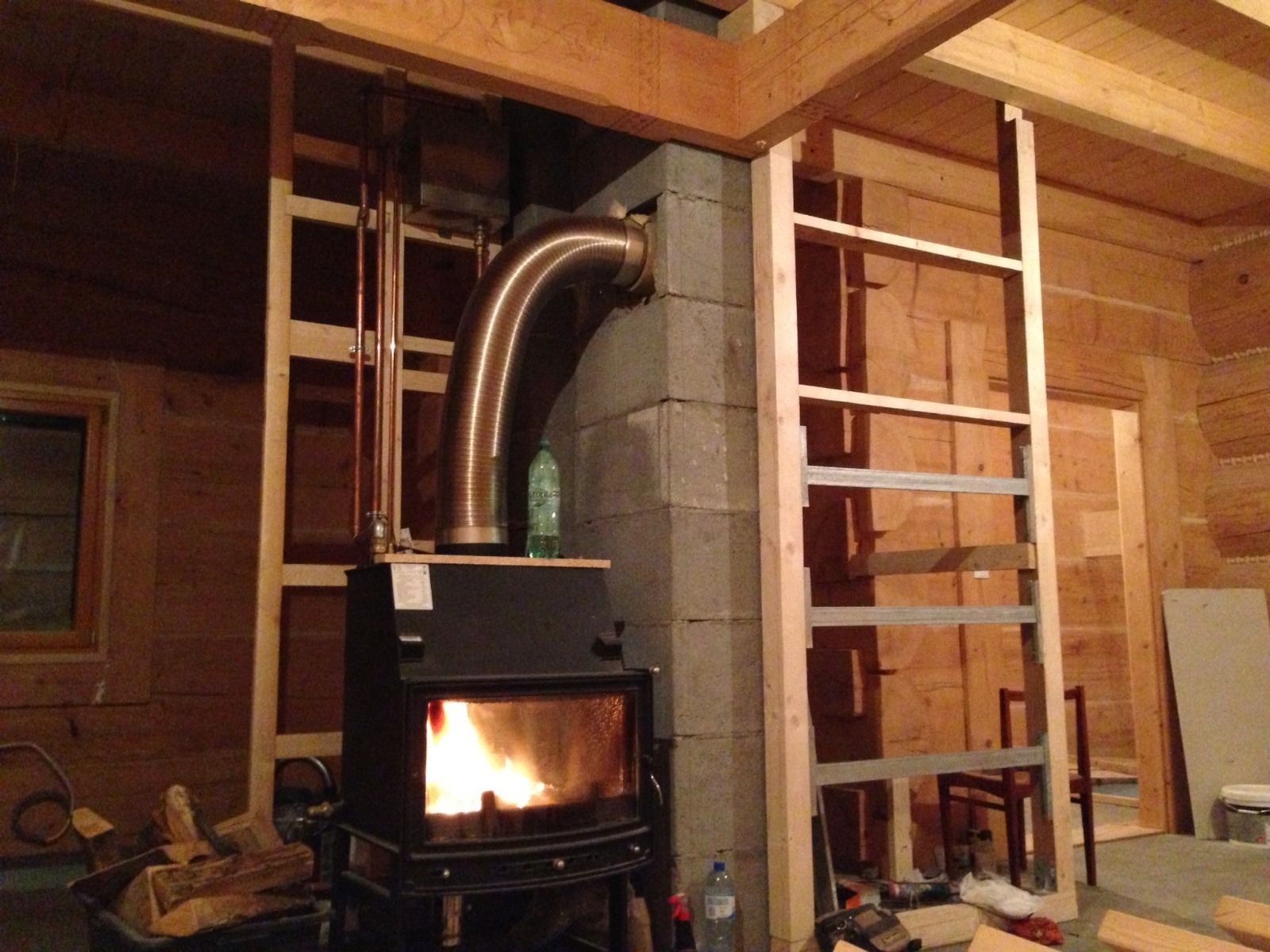 Wnętrze drewnianego domu w trakcie budowy kominka z rurą odprowadzającą dym. Widoczne elementy instalacji i narzędzia.