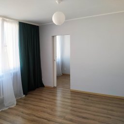 NDF Zbigniew Czerwień - Pomalowane na szaro wnętrze pokoju z drewnianą podłogą, białymi zasłonami i ciemnozielonymi kotarami, widoczny okrągły, papierowy klosz lampy sufitowej i otwarte drzwi do sąsiedniego pomieszczenia.