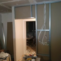 NDF Zbigniew Czerwień - Wnętrze w trakcie prac: montaż stelaża z płyt gipsowo-kartonowych, widoczne przewody elektryczne, narzędzia i materiały budowlane na podłodze.