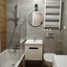 Nowoczesna łazienka w szarościach z drewnopodobnymi akcentami: wanna z parawanem, umywalka z szafką, okrągłe lustro, grzejnik drabinkowy i wisząca toaleta.