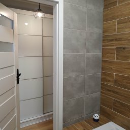 NDF Zbigniew Czerwień - Wykończona łazienka z szarymi i drewnopodobnymi płytkami, białą wanną i otwartymi drzwiami do garderoby z przesuwnymi drzwiami, widoczna lampa sufitowa w stylu industrialnym.